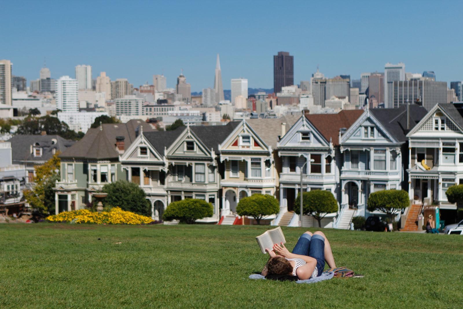 A woman lounges on a blanket in a park adjacent to Victorian Row Houses in San Francisco.